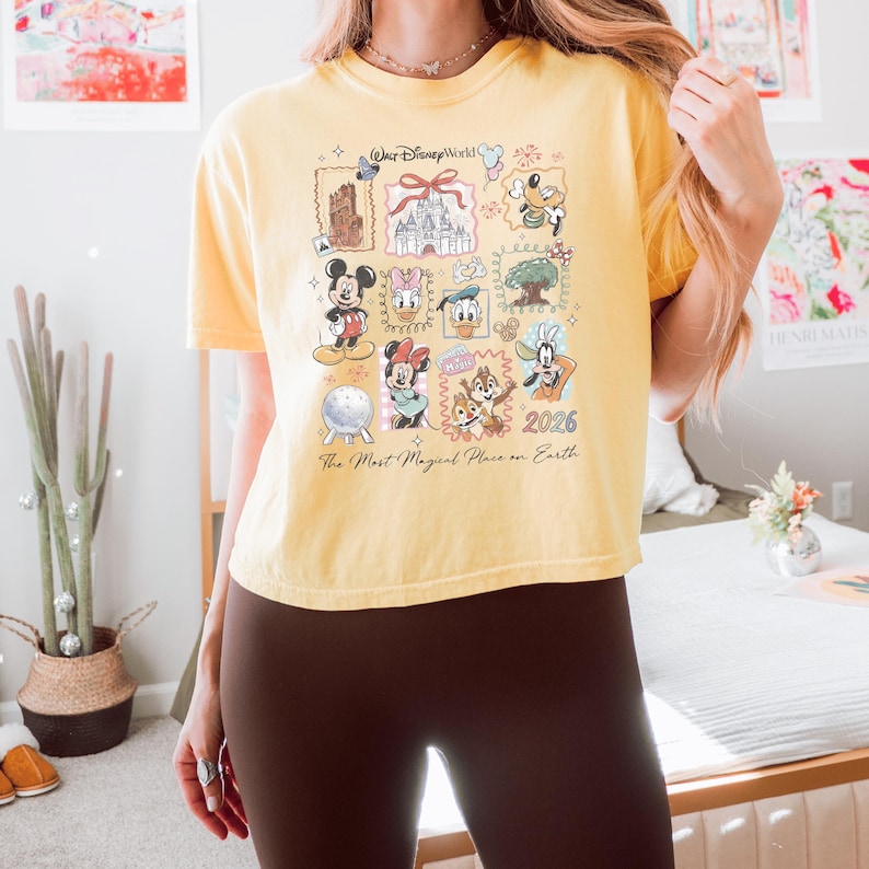K&ouml;nnte beinhalten: Ein blassgelbes Kurzarm-T-Shirt mit einem Grafikdruck von Walt Disney World-Figuren und Wahrzeichen im Briefmarken-Layout. Das Design umfasst Mickey, Minnie, Donald, Daisy, Goofy, Pluto, Chip, Dale sowie den Text 2026.