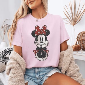 Pode incluir: Uma t-shirt de manga curta em rosa claro com uma estampa gr&aacute;fica desgastada da Minnie Mouse. A personagem &eacute; retratada com as m&atilde;os sob o queixo, usando o seu la&ccedil;o vermelho caracter&iacute;stico com bolinhas brancas e uma saia a combinar.