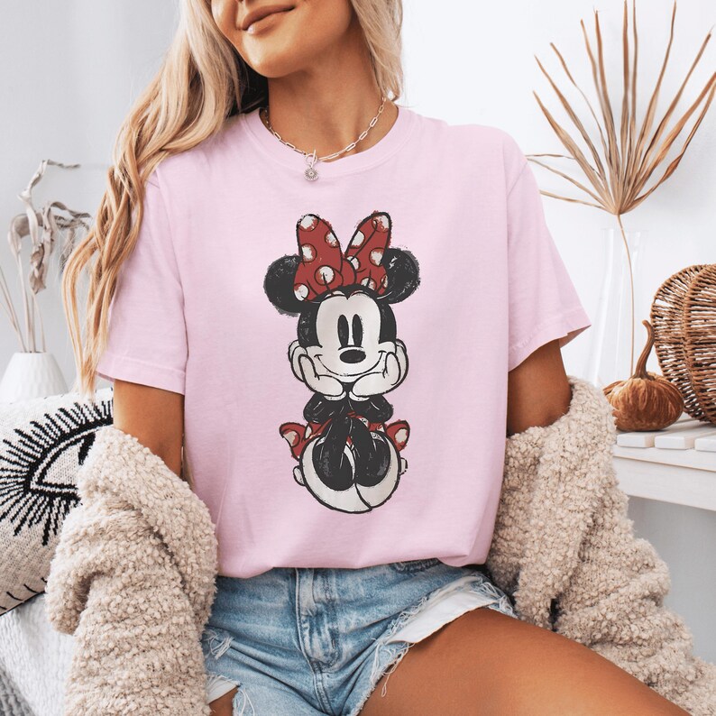 Pode incluir: Uma t-shirt de manga curta em rosa claro com uma estampa gr&aacute;fica desgastada da Minnie Mouse. A personagem &eacute; retratada com as m&atilde;os sob o queixo, usando o seu la&ccedil;o vermelho caracter&iacute;stico com bolinhas brancas e uma saia a combinar.