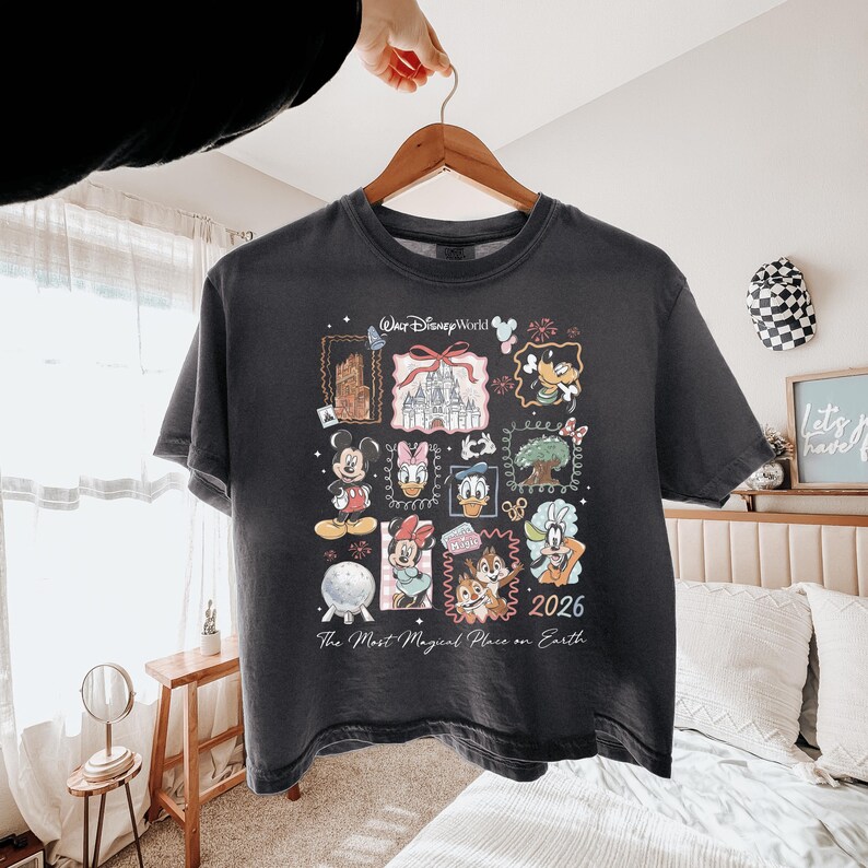 K&ouml;nnte beinhalten: Ein anthrazitfarbenes Kurzarm-T-Shirt mit einem Grafikdruck von Walt Disney World, der Illustrationen von Mickey Maus, Minnie Maus, Donald Duck, Daisy Duck, Goofy, Chip, Chap und den Text The Most Magical Place on Earth 2026 zeigt.