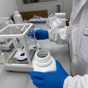 May include: A laboratory analytical balance with a glass draft shield enclosure, featuring a white plastic bottle held by blue gloved hands. The scale displays a small amount of dark blue powder on a white weighing paper inside the chamber.