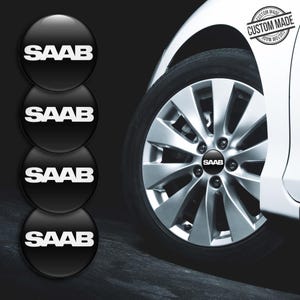 May include: A set of four circular black wheel centre caps featuring the white SAAB logo. One cap is installed on a silver multi-spoke car wheel, while three identical caps are stacked vertically to the left. A custom made stamp is visible.