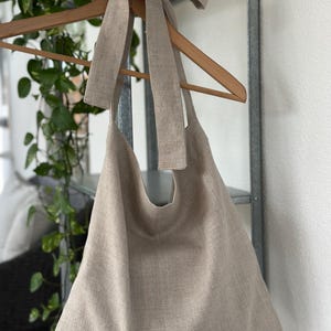 May include: A large, beige linen tote bag with a long linen strap tied into a bow at the top. The bag is hanging on a wooden hanger.