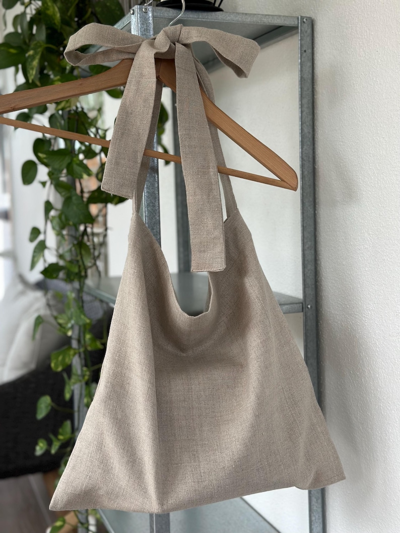 May include: A large, beige linen tote bag with a long linen strap tied into a bow at the top. The bag is hanging on a wooden hanger.