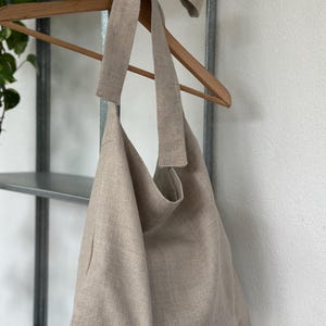 May include: A beige linen tote bag with a long, knotted strap. The bag is hanging on a wooden hanger.