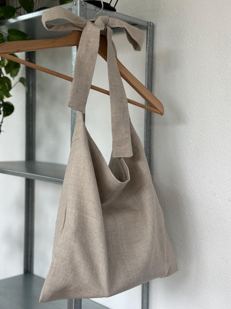May include: A beige linen tote bag with a long, knotted strap. The bag is hanging on a wooden hanger.