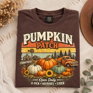 May include: A brown short-sleeve t-shirt featuring a graphic of a pumpkin patch with a truck, barns, and sunflowers. The text reads PUMPKIN PATCH, FARM FRESH, and Open Daily U-PICK HAYRIDES CIDER. The design includes orange, yellow, and green colors.