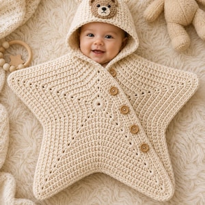Könnte beinhalten: Eine cremefarbene, sternförmige Häkeldecke für Babys mit einer Kapuze, die mit einem kleinen Bärengesicht und Bärenohren verziert ist. Die Decke hat eine diagonale Öffnung, die mit fünf hellbraunen Holzknöpfen verschlossen wird.