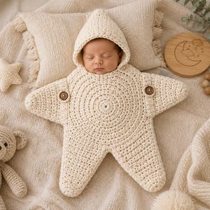Op de afbeelding: Een crèmekleurige, stervormige gehaakte slaapzak met capuchon, voorzien van een cirkelvormig steekpatroon in het midden en twee bruine knopen op de bovenarmen. Het kledingstuk is ontworpen om een baby te omhullen met een zachte textuur.