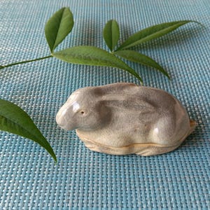 May include: A small, smooth, ceramic rabbit figurine in a resting position with a glossy, mottled grey and beige finish. The rabbit has subtle sculpted ears laid back against its body and a rounded form, presented as a singular decorative object.