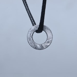May include: A circular pendant with a metallic, silver-toned surface featuring a distinct cross-hatched, crystalline pattern. The pendant is suspended from a thin, black braided cord that passes through the centre opening of the ring.