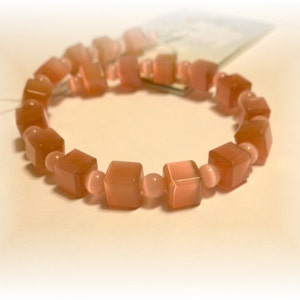 May include: A bracelet featuring alternating cube and round beads in shades of peach and light pink. The cube beads have a translucent quality, and the bracelet is strung on a flexible cord. This jewelry piece is suitable for everyday wear.