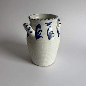 Può includere: Un vaso in ceramica color crema con due manici laterali, caratterizzato da motivi a pennellate blu dipinti a mano attorno al bordo e alla parte superiore. La superficie esterna presenta striature orizzontali visibili e una finitura lucida.