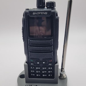May include: A black Baofeng two-way radio with a digital screen, keypad, and antenna, resting in a grey plastic stand featuring the text IsTriDesigns. The radio has a tall, thin, silver telescopic antenna attached to the side of the base.