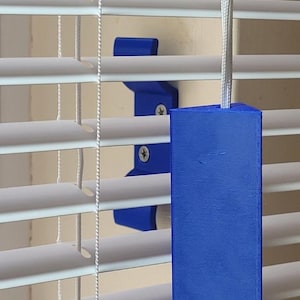 May include: A blue plastic blind cord cleat and a matching blue rectangular cord pull. The cleat is mounted to the wall with two silver screws, while the cord pull hangs vertically from the white cord of the horizontal window blinds.