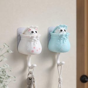 Puede incluir: Dos ganchos adhesivos de pared que incluyen una bolsa blanca con detalles de flores rosas y una bolsa azul claro. Cada bolsa contiene una pequeña figura de gato blanco con ojos y bigotes negros. Un llavero plateado cuelga del gancho blanco.