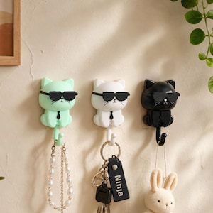 Puede incluir: Tres ganchos de pared con forma de gatos que llevan gafas de sol negras y corbatas. Los gatos son de color verde claro, blanco y negro. Cada gato tiene una cola que funciona como gancho, sosteniendo una pulsera de perlas, un llavero con una etiqueta Ninja y un peluche.