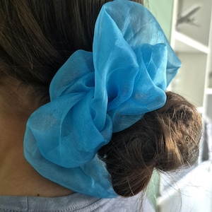 May include: A large, bright blue hair scrunchie made of a sheer, translucent fabric. The accessory is gathered into voluminous, ruffled folds, creating a textured and layered appearance while secured around a bun of dark brown hair.