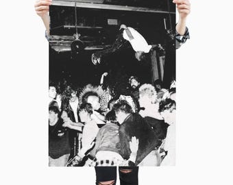 May include: A black and white photographic print depicting a crowded indoor concert scene. A person in a white t-shirt is captured mid-air performing a backflip above a dense crowd of people with various hairstyles, including mohawks and long hair.
