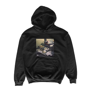 May include: A black hooded sweatshirt featuring a square graphic print on the chest. The print depicts a cluttered desk surface with various objects, including a small white cup, gold-colored items, and papers with the number 80 visible.