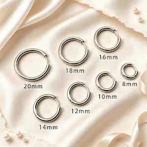 Może przedstawiać: Kolekcja siedmiu srebrzystych kolczyków w kształcie kół, ułożonych według rozmiaru i oznaczonych wymiarami 8mm, 10mm, 12mm, 14mm, 16mm, 18mm oraz 20mm. Każdy kolczyk ma prosty, okrągły kształt z mechanizmem zamykającym na zawiasie.