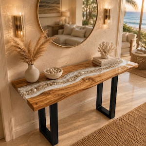 May include: A rectangular console table featuring a natural wood slab top with a central river-like inlay of white resin and smooth river stones. The table is supported by two black metal U-shaped legs and holds a vase with pampas grass and a bowl.