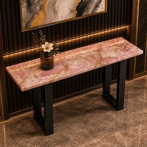 Puede incluir: Una mesa consola rectangular con una superficie de piedra rosa pulida con vetas naturales blancas y doradas, sostenida por dos patas de metal negro en forma de U. Un jarrón de vidrio transparente con una flor blanca descansa sobre la mesa.