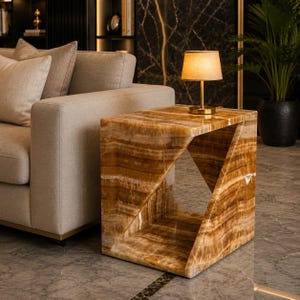 May include: A square side table crafted from polished amber-colored onyx stone with a distinct geometric cutout design. The table features horizontal banding patterns throughout the natural stone surface and supports a small brass lamp with a beige shade.