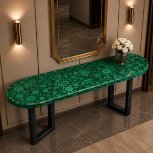 Puede incluir: Una mesa consola ovalada y alargada con un tablero de patrón de malaquita verde vibrante con detalles circulares concéntricos. La mesa se apoya en dos patas de metal negro en forma de U, proporcionando una base estable para la superficie pulida.