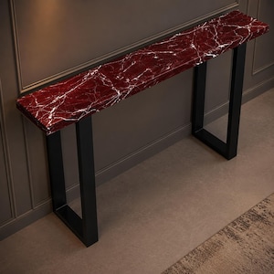 Peut inclure: Une console longue et étroite dotée d'un plateau rectangulaire en marbre rouge profond, marqué par des veines blanches prononcées. La table est soutenue par deux pieds en métal noir robustes en forme de U, placés aux extrémités de la structure.