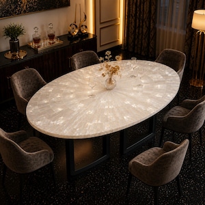 May include: An oval dining table with a white, iridescent mother-of-pearl tiled surface and a black metal base, surrounded by six dark grey upholstered armchairs. A glass vase with gold-toned decorative branches sits in the centre.
