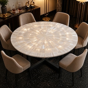 May include: A round dining table featuring a sunburst pattern of iridescent white tiles on the tabletop, supported by a black metal cross-base. Six upholstered chairs with vertical channel tufting in a light beige fabric surround the table.