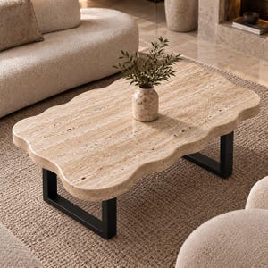 Puede incluir: Una mesa de centro con un tablero de piedra travertino beige de bordes ondulados y una estructura de metal negro. Un pequeño jarrón de travertino beige a juego con una planta de hojas verdes se sitúa en el centro de la superficie de piedra natural.