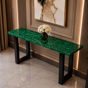 Puede incluir: Una mesa consola rectangular con un tablero de patrón de malaquita verde vibrante y acabado pulido. La mesa se apoya sobre dos patas robustas de metal negro en forma de U. Un jarrón de vidrio oscuro con un ramo de flores blancas se sitúa encima.