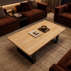 May include: A rectangular coffee table with a beige travertine stone top and black metal U-shaped legs. A book titled Picasso and a small metallic bowl are placed on the surface. The table features a natural porous texture and rounded edges.
