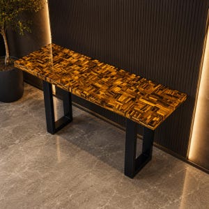 May include: A rectangular console table featuring a polished tiger's eye gemstone surface with a mosaic pattern of golden-brown and black tones. The table is supported by two sturdy, matte black metal U-shaped legs that provide a stable base.
