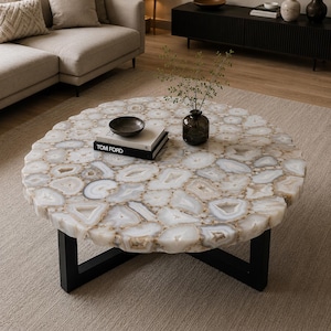 May include: A round coffee table featuring a tabletop constructed from numerous translucent white and grey agate slices arranged in a mosaic pattern, supported by a black metal base. A small dark glass vase and a book titled TOM FORD sit on top.