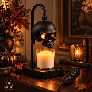May include: A black metal candle warmer lamp featuring a skull-shaped top that emits a warm glow, positioned above a glass jar candle. The device includes a rectangular base and a corded remote control with power, timer, and brightness settings.