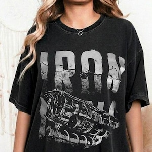 May include: A black short-sleeve t-shirt featuring a distressed white graphic print of the word IRON and a detailed mechanical hand illustration. The garment has a crew neckline and a relaxed, oversized fit with a faded, vintage-inspired aesthetic.