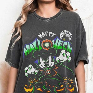 May include: A charcoal gray short-sleeve t-shirt featuring a graphic of a cartoon mouse dressed as a vampire with green claws and a cape. The text reads Happy Halloween in green and white letters, surrounded by orange jack-o'-lanterns and bats.
