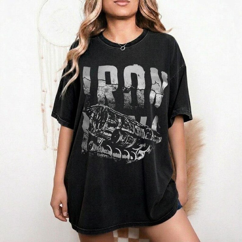 May include: A black short-sleeve t-shirt featuring a distressed white graphic print of the word IRON and a detailed mechanical hand illustration. The garment has a crew neckline and a relaxed, oversized fit with a faded, vintage-inspired aesthetic.