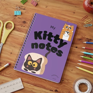 May include: A purple spiral-bound notebook with the text My Kitty notes printed in black. The cover features a cartoon orange cat holding a white mug and a black cat face inside a slice of bread. The notebook is surrounded by office supplies.