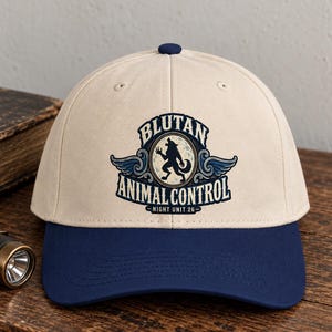 Puede incluir: Una gorra de béisbol beige con visera azul oscuro y un botón superior. La parte frontal presenta un gráfico circular de una silueta de hombre lobo, alas azules y el texto BLUTAN ANIMAL CONTROL NIGHT UNIT 26 en letras azul oscuro.