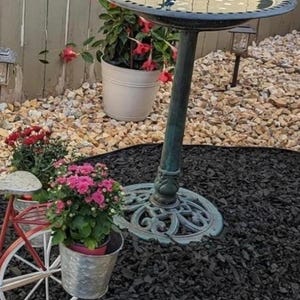 May include: A dark green metal bird bath with a circular basin and an ornate pedestal base, positioned near a white metal bicycle-shaped plant stand holding a silver bucket filled with pink flowers and a white pot with red flowers.