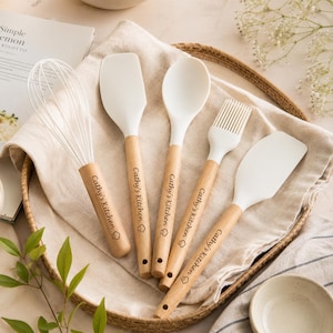 Può includere: Un set di cinque utensili da cucina con teste in silicone bianco e manici in legno chiaro. Ogni manico è inciso con il testo 'Cathy's Kitchen' e una piccola icona di un cappello da chef. Il set include una frusta, due spatole, un cucchiaio e un pennello.