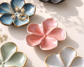 May include: Five ceramic flower-shaped dishes in blue, black, pink, light green, and white. The blue dish holds a gold necklace with a pendant, two pearl stud earrings, and a gold ring. Each dish features five rounded petals and a glazed finish.
