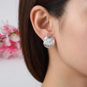 May include: A silver stud earring shaped like a seashell with a polished finish and vertical ridges. The earring is circular in form and features a scalloped edge, reflecting light across its metallic surface while attached to an earlobe.