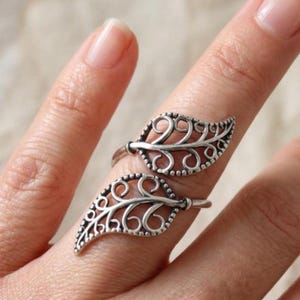 May include: A silver-toned ring featuring two openwork leaf designs with intricate swirling filigree patterns. The metal has a darkened, oxidized finish within the recessed areas to highlight the detailed scrollwork of the leaf-shaped motifs.