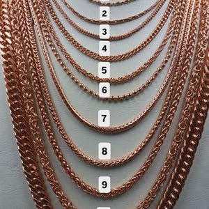 May include: A collection of eleven rose gold-toned metal chains displayed on a neck form, numbered 1 through 11. The chains vary in thickness and link style, ranging from thin, delicate cable and bead chains to thicker, heavy curb and woven link designs.