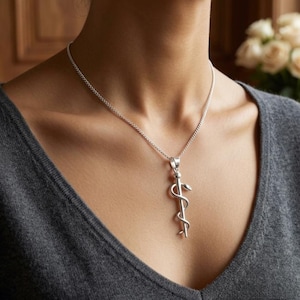 Puede incluir: Un collar de tono plateado con un colgante en forma de Vara de Esculapio, que consiste en un bastón vertical entrelazado por una serpiente. El colgante cuelga de una delicada cadena de tono plateado sobre una tela gris texturizada.